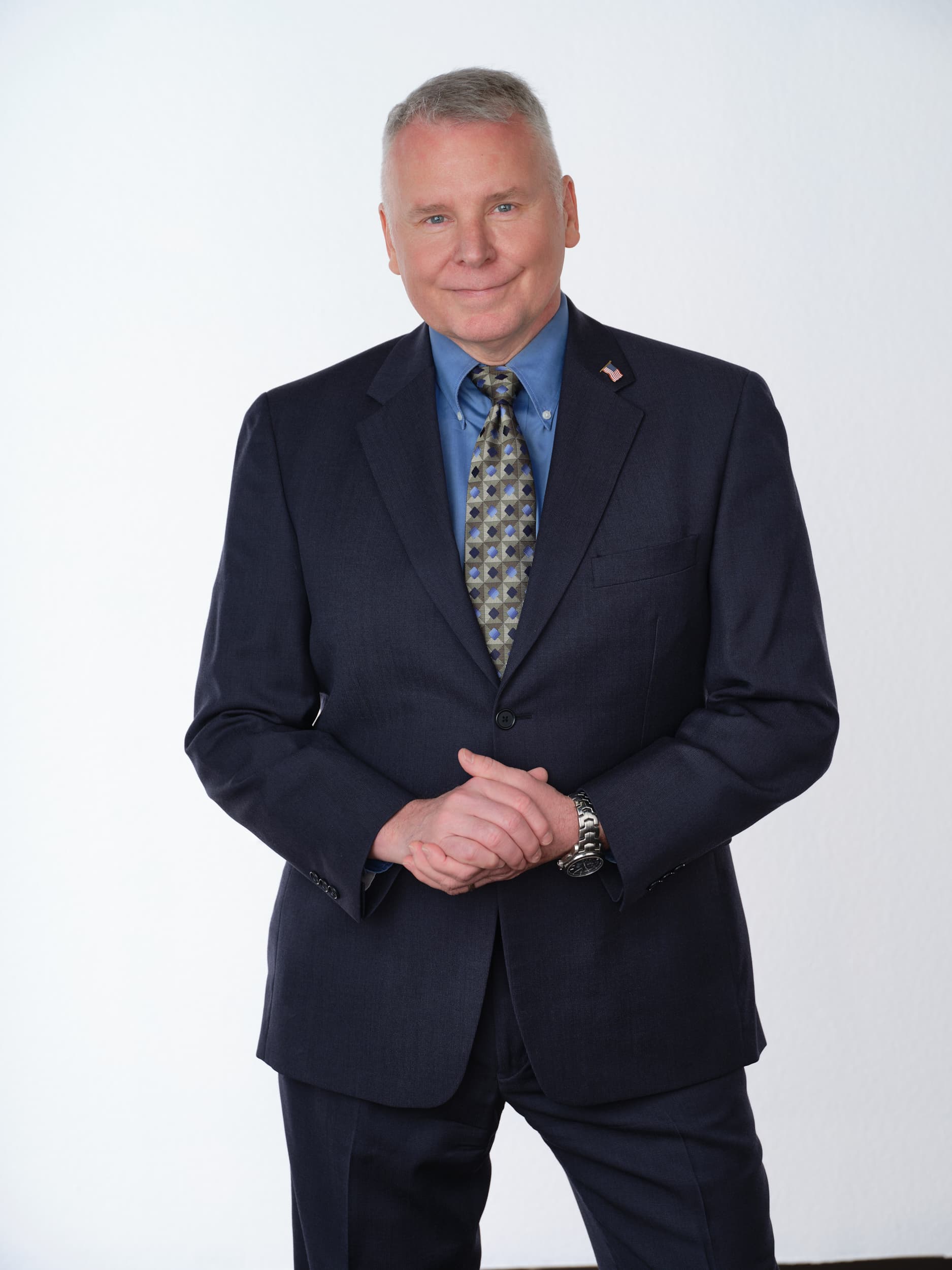 Smiling man in a navy suit and patterned tie, hands clasped, against a white background.
