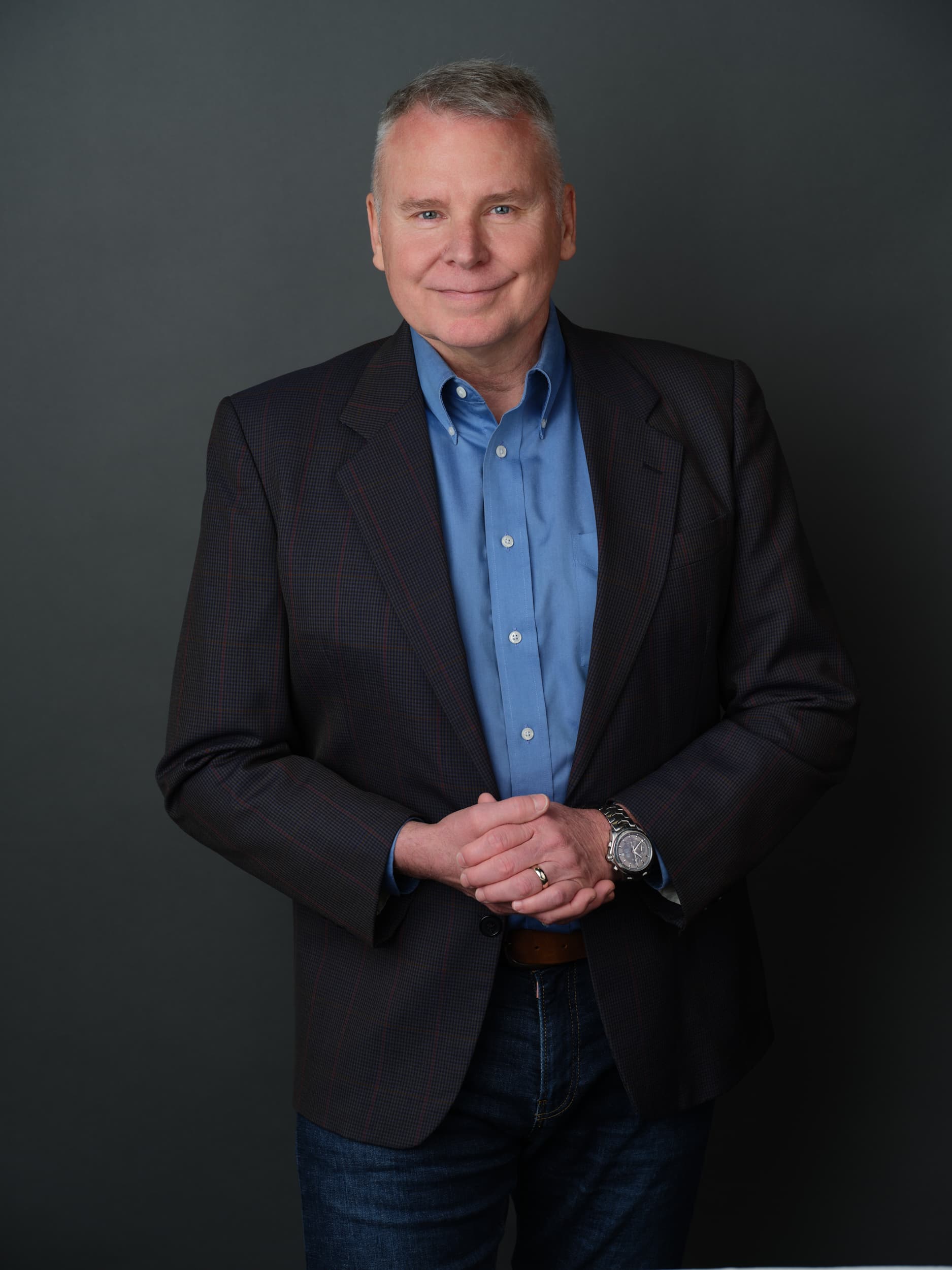 Smiling man in a blue shirt and dark blazer with hands clasped against gray background.
